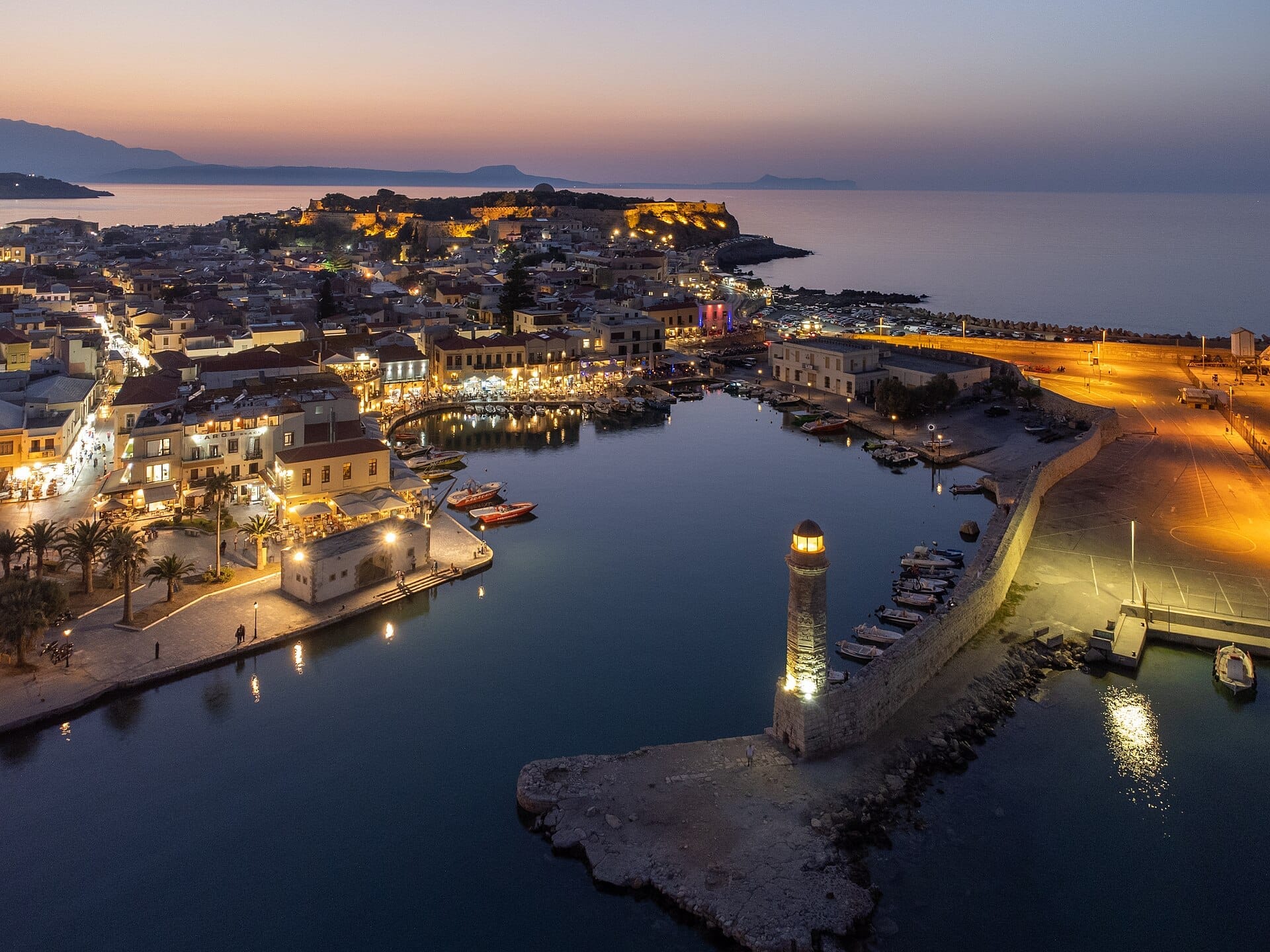 Vista aerea della città vecchia di Rethymno al crepuscolo con il porto veneziano illuminato, il faro in pietra, i caffè sul lungomare e il forte medievale sullo sfondo.