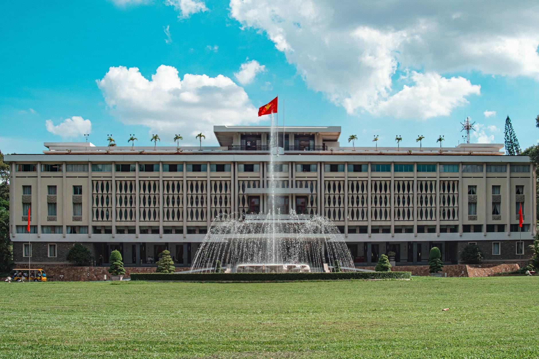 Vista amplia y simétrica del Palacio de la Reunificación en Ciudad Ho Chi Minh, con una gran fuente y la bandera vietnamita bajo un cielo azul intenso.