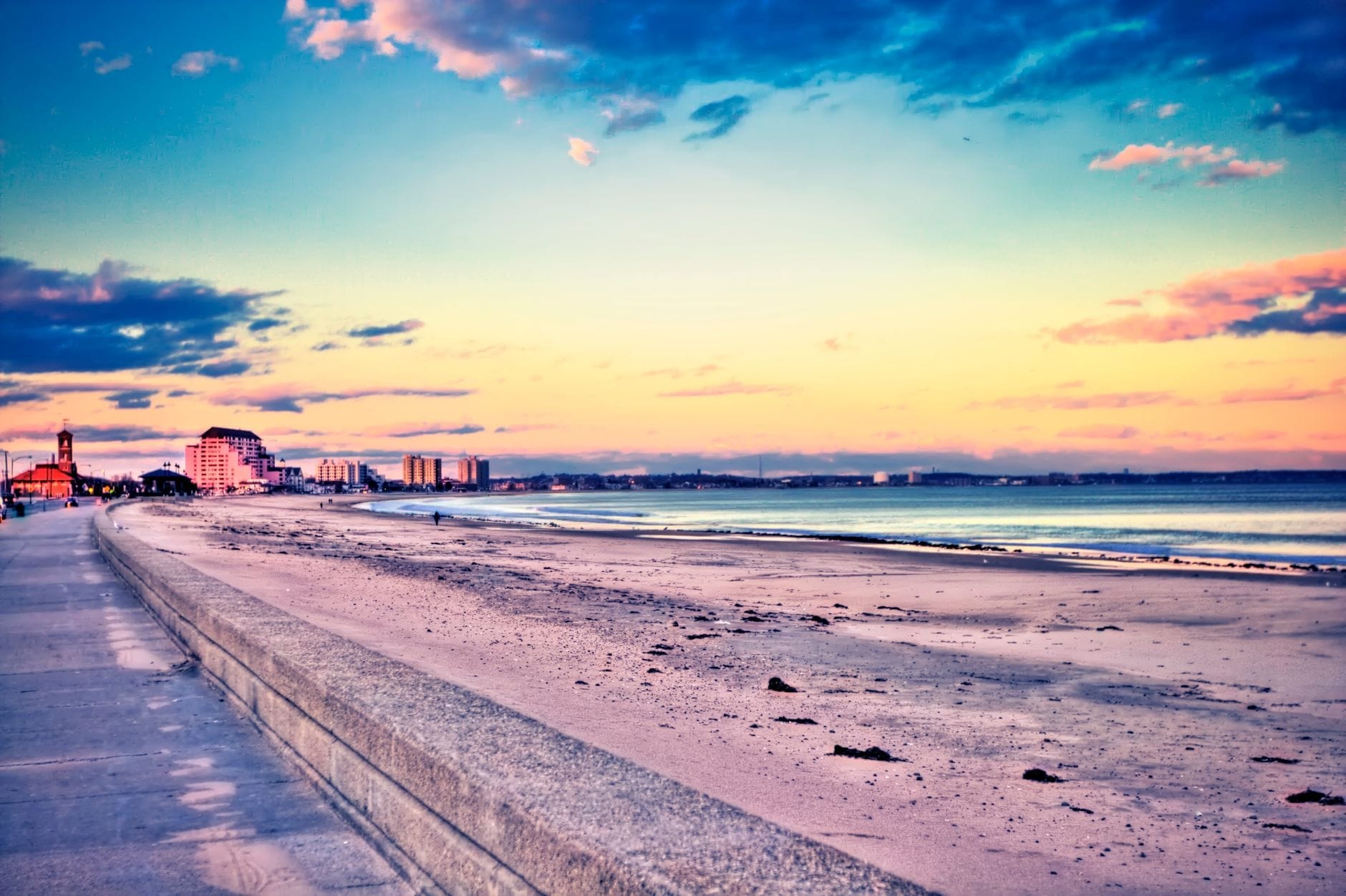 Una lunga distesa di sabbia chiara affiancata da un ampio lungomare, con le onde dell'oceano e gli edifici della zona di Boston che si stagliano in lontananza sotto un cielo colorato al tramonto.