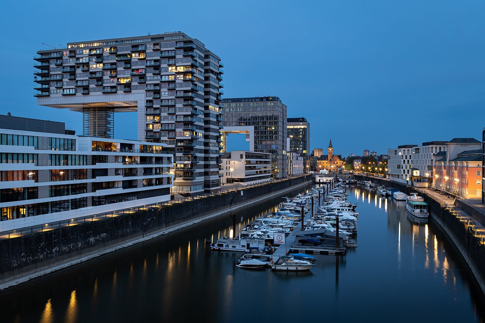 ภาพมุมเย็นของย่าน Rheinauhafen ในโคโลญจน์ เห็นตึก Kranhäuser ส่องแสง อาคารริมน้ำยุคใหม่ และท่าจอดเรือที่มีเรือจอดแน่นท่าภายใต้ท้องฟ้าสีคราม
