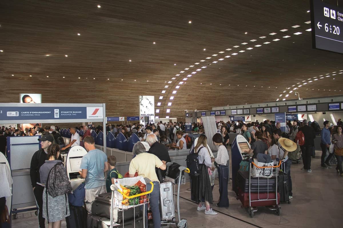 Busy airport check-in area with crowds of travelers and luggage carts in a modern terminal with wood-patterned ceiling.