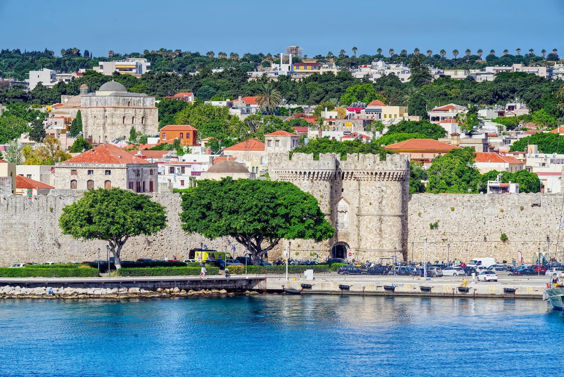 Vista vibrante da cidade de Rodes com muralhas medievais de pedra, edifícios históricos, árvores frondosas e um mar azul intenso ao fundo sob um céu limpo.