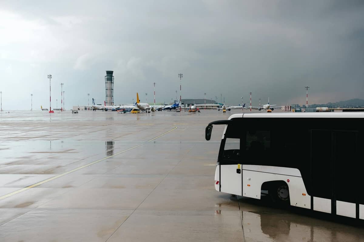 A white airport bus on the tarmac with planes and a control tower visible in the background under a cloudy sky.