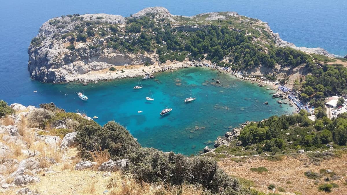 Aerial view of a scenic turquoise bay in Rhodes with anchored boats, surrounded by rocky cliffs and greenery.