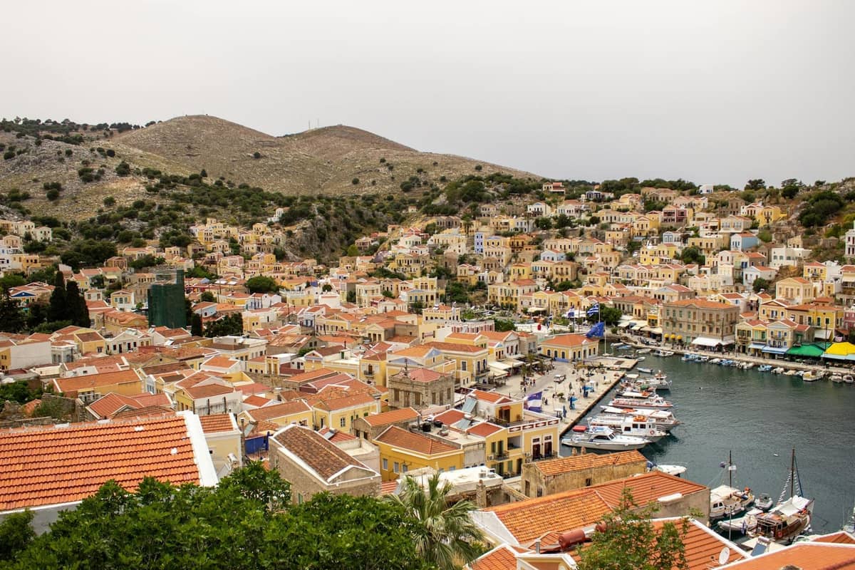 Overhead view of the colorful neoclassical harbor and hillside houses of Symi Island, boats moored along the waterfront, under a cloudy sky.