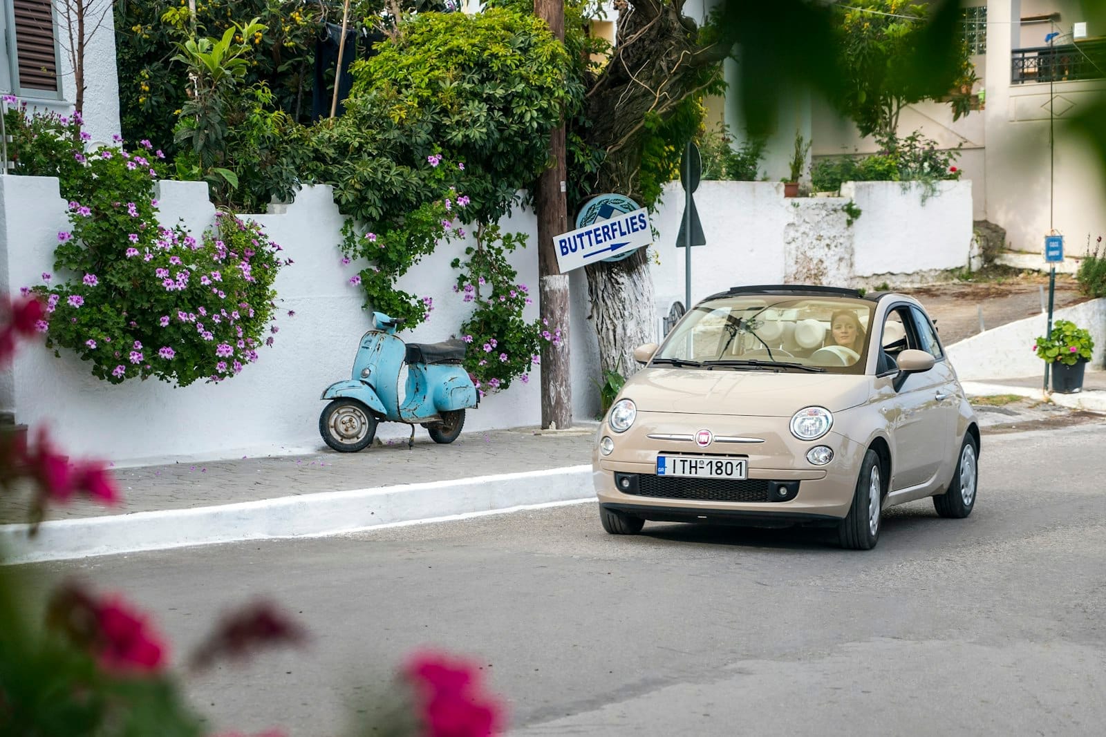 Um Fiat 500 bege passa por uma parede caiada com flores cor-de-rosa, uma scooter azul vintage e uma placa com a inscrição 'Butterflies' em Rodes.