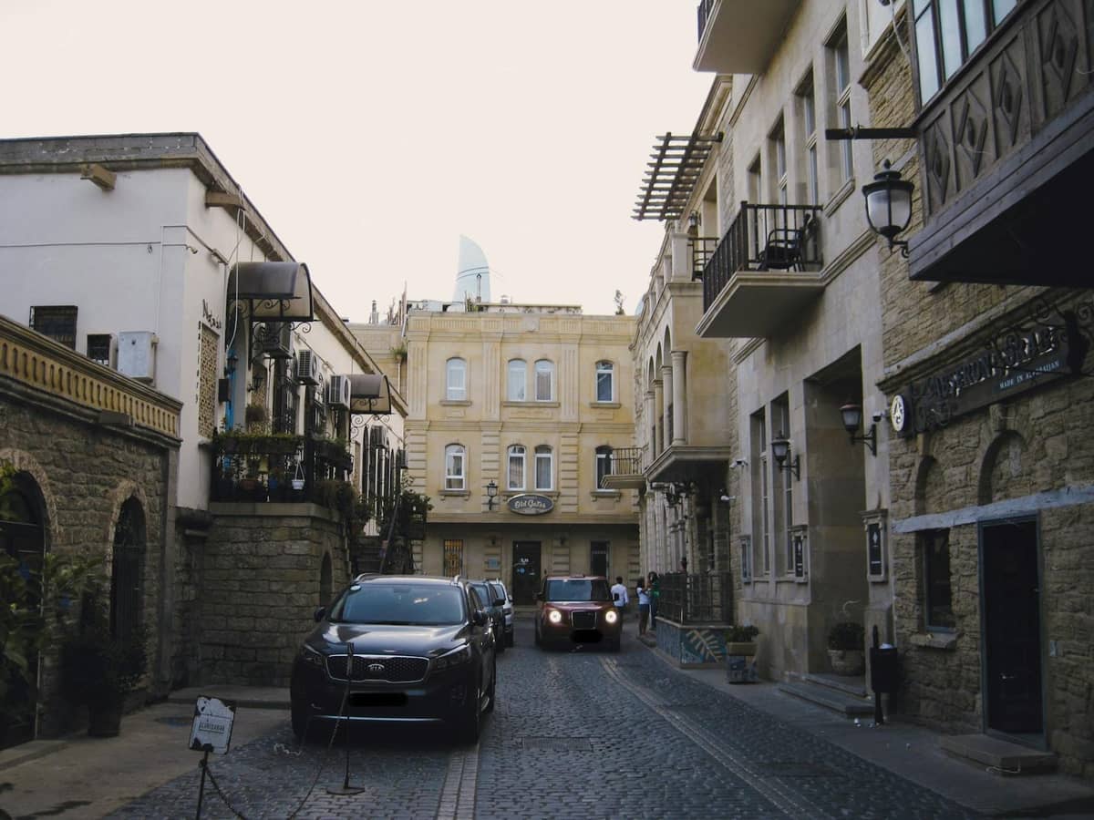 Cars parked along a narrow cobblestone street lined with historic Mediterranean-style buildings in a town setting.