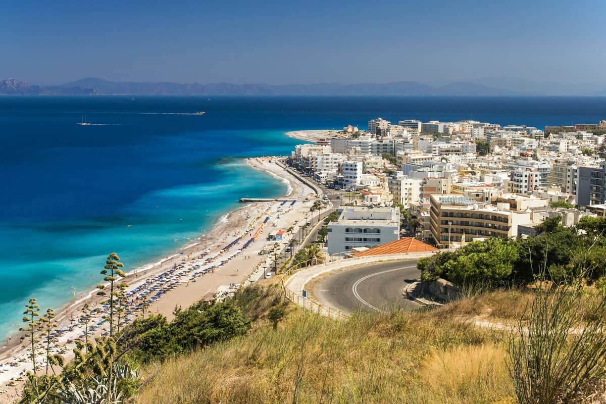 Coastal cityscape of Rhodes with winding road, beachfront, and rows of white buildings along a vibrant blue sea on a sunny day.