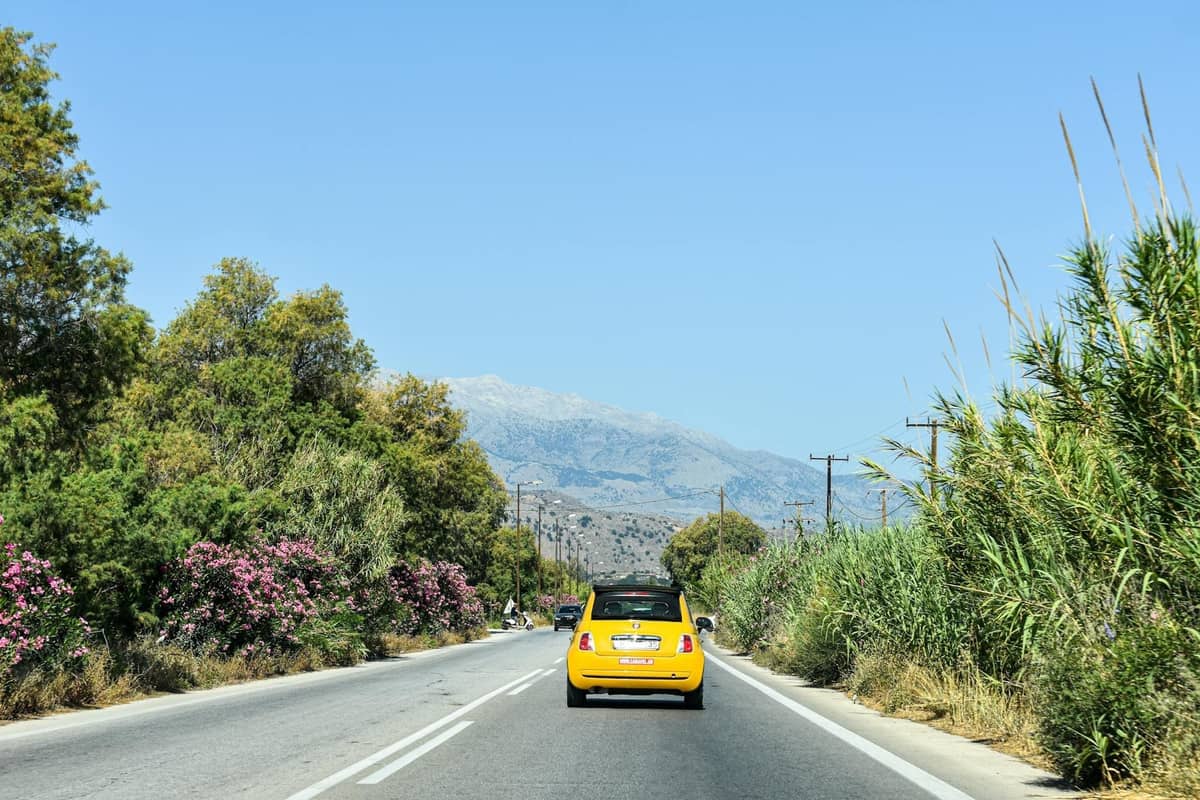 A well-maintained road in Rhodes with a yellow car driving, surrounded by greenery and distant mountains under a clear blue sky.