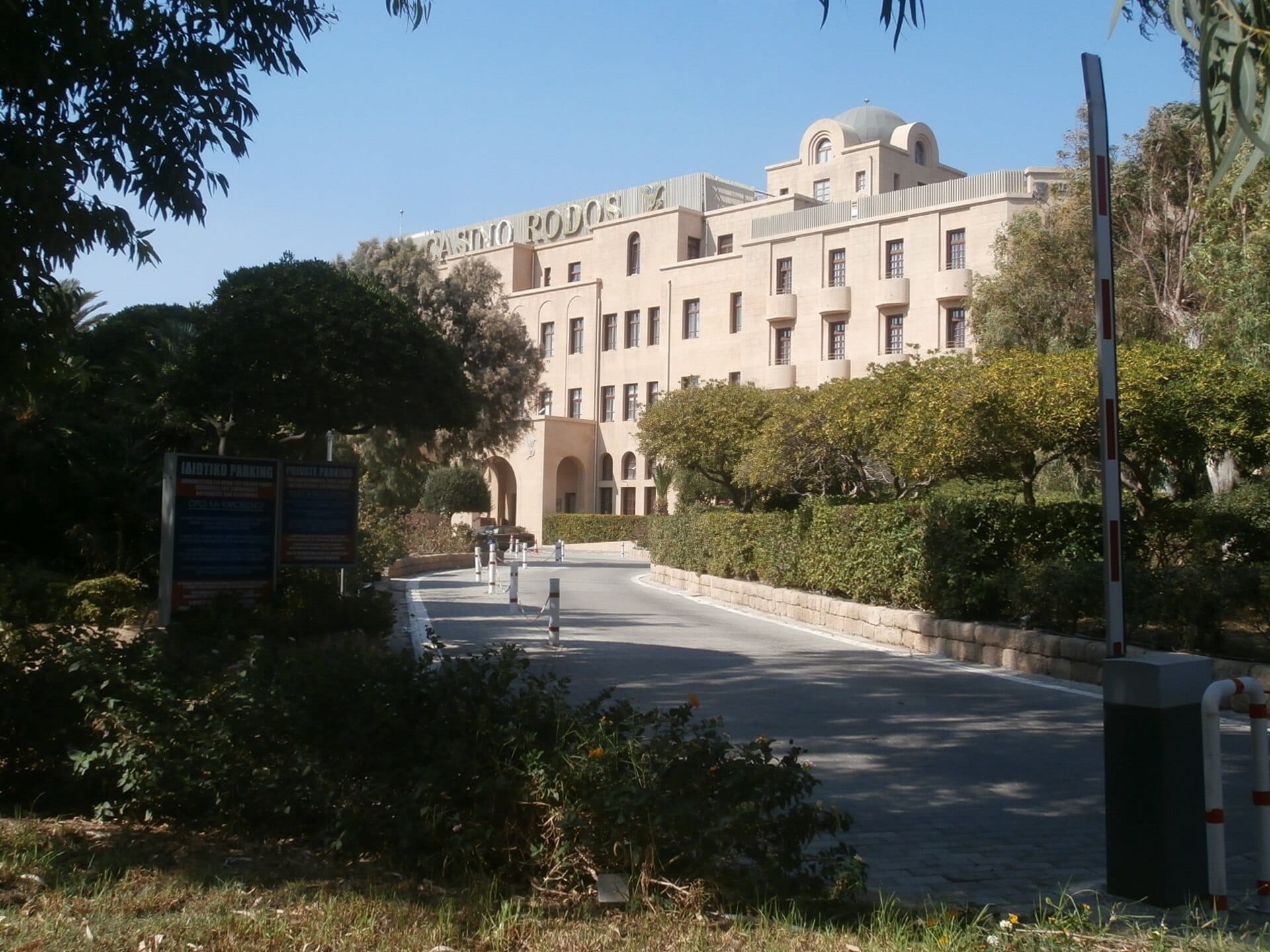 Grande Albergo delle Rose ที่ตั้งของ Casino Rodos ถ่ายในแสงแดดพร้อมด้านหน้าอาคารอันสง่างามและทางเข้าที่เรียงรายด้วยต้นไม้ในเมืองใหม่ของ Rhodes