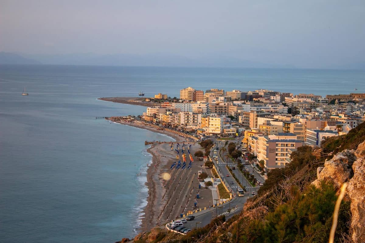 Elevated view of Rhodes coastline and city, with buildings stretching along the shore and the sea at sunset, seen from a high vantage point.