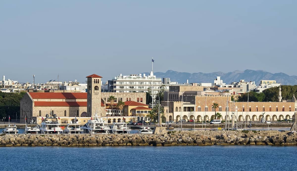 View of Mandraki Harbour waterfront featuring Italianate government buildings and a prominent clocktower, with yachts docked along the promenade and hills in the background.