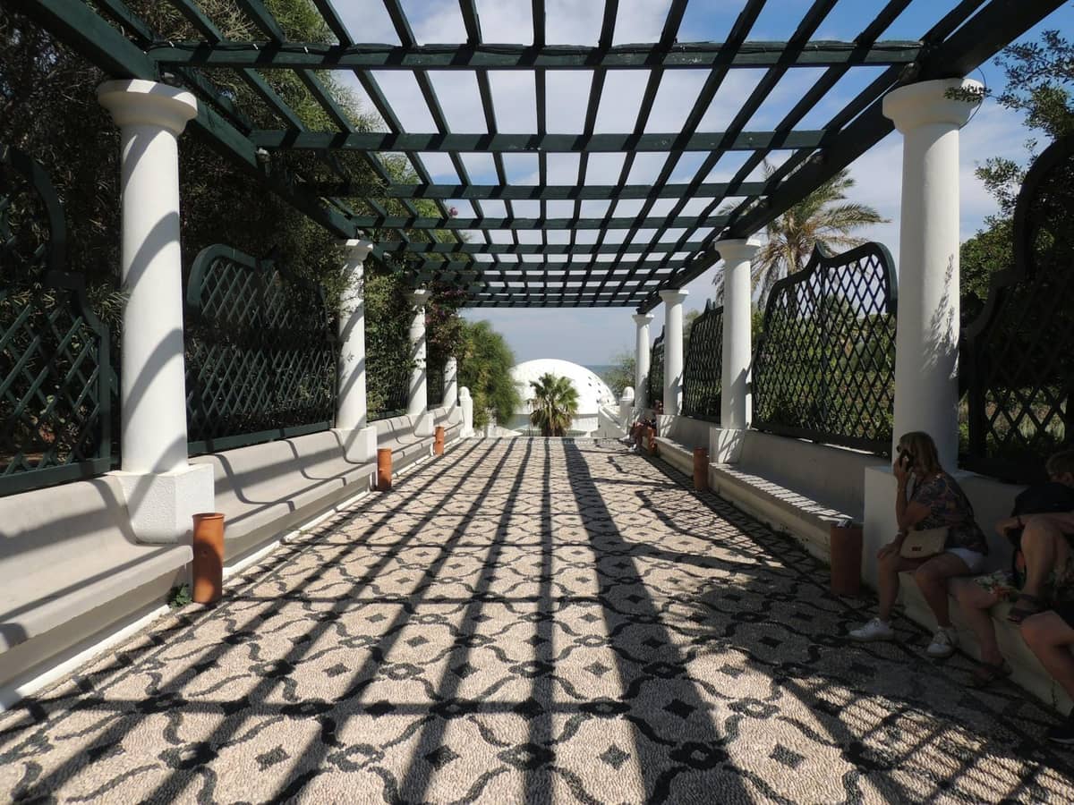 Colonnaded outdoor walkway with mosaic stone floor and lattice overhead, people sitting along the sides, lush greenery outside.