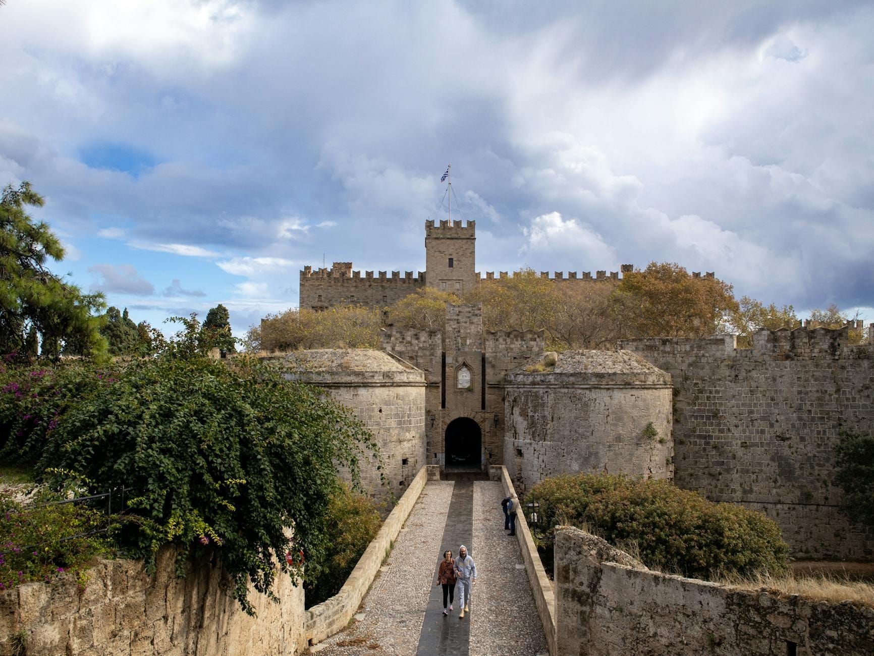 Muralhas e entrada de um castelo medieval de pedra com passarela cercada por vegetação exuberante e céu dramático em Rodes, Grécia, transmitindo uma sensação de história oculta.