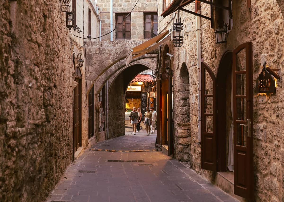 Stone-paved narrow alley in Rhodes’ old town with warm lighting and an arched passageway, featuring rustic stone buildings and a couple walking together.