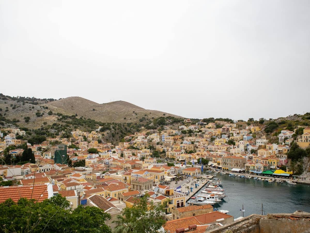 Sweeping view of a harbor town with colorful houses, boats docked along the waterfront, and dry hills rising in the background.