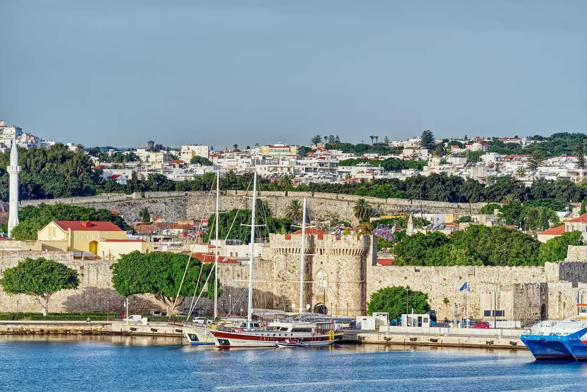 Harbor scene in Rhodes with medieval city walls, ferries, sailboats, and the old town visible in the background on a clear day.