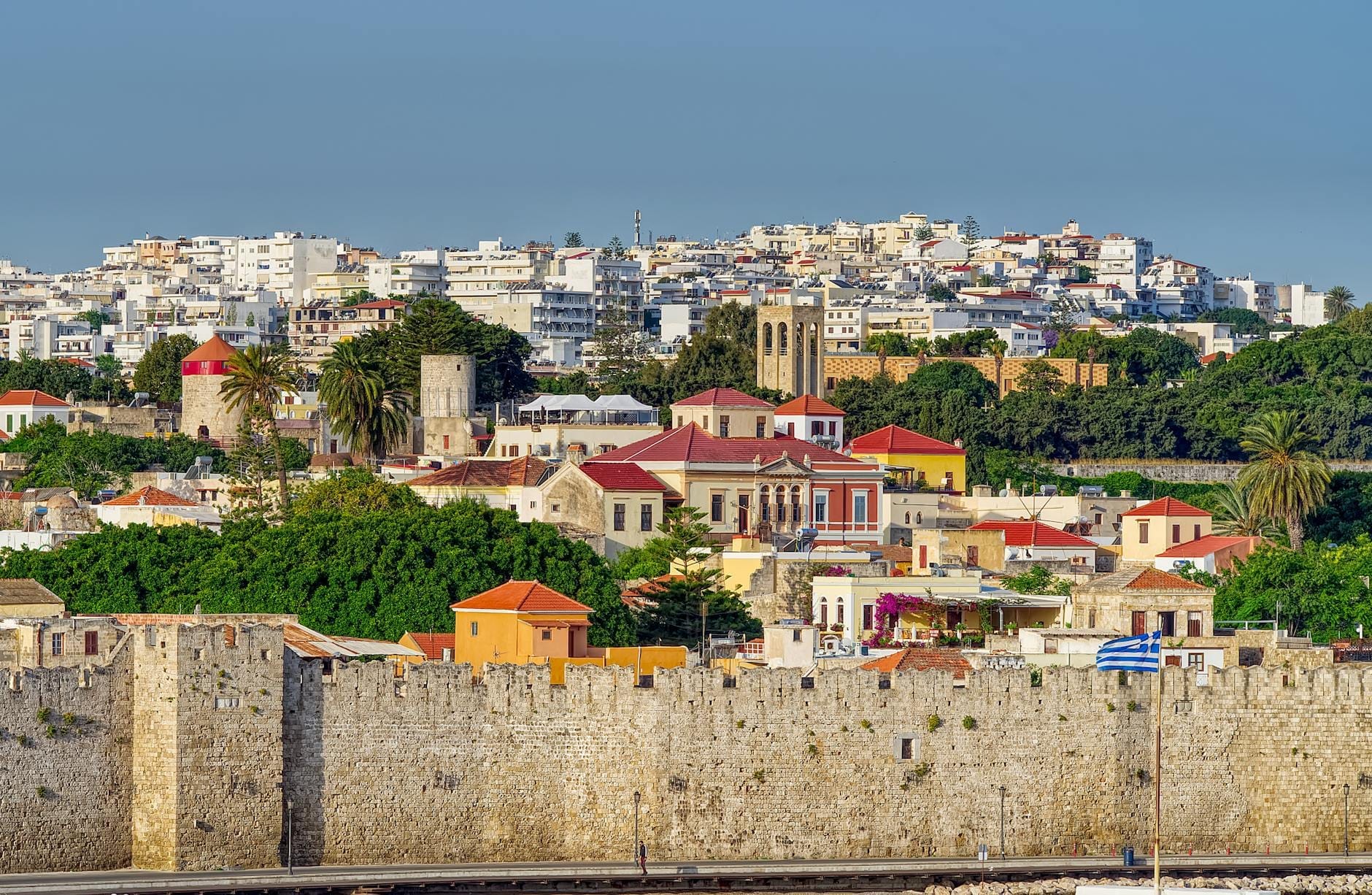 Vista das muralhas medievais da Cidade Velha de Rodes, casas coloridas, árvores verdes exuberantes e edifícios modernos sob céus limpos de outubro.