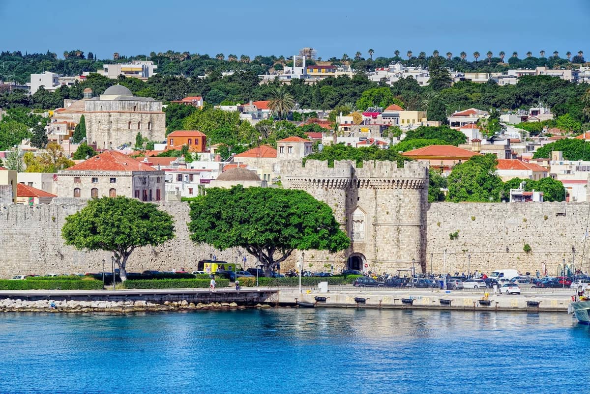 View of Rhodes old town with stone walls, historic buildings, lush trees, and clear blue sky by the waterfront under sunny weather.