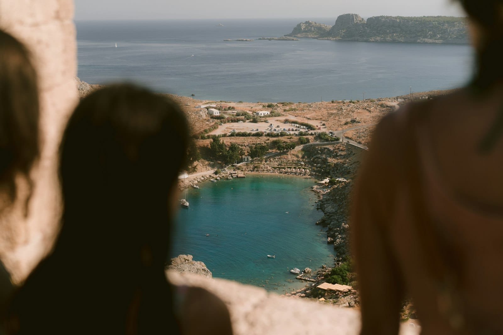 Vista aérea da baía de Lindos em Rodes com águas turquesas, guarda-sóis na praia, costa rochosa e promontórios ao longe.