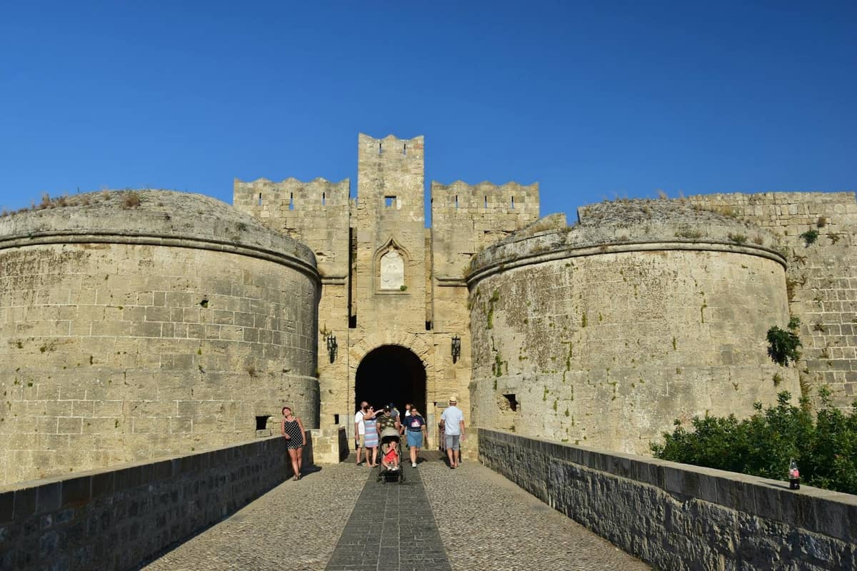 Tourists walking through a large medieval stone gate in Rhodes under clear blue summer skies.