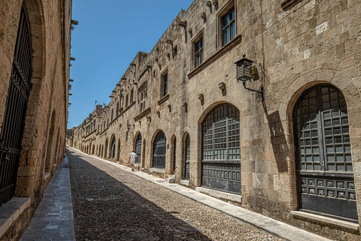 A wide cobblestone street bordered by imposing medieval stone buildings with arched doorways and shuttered windows under a bright blue sky.