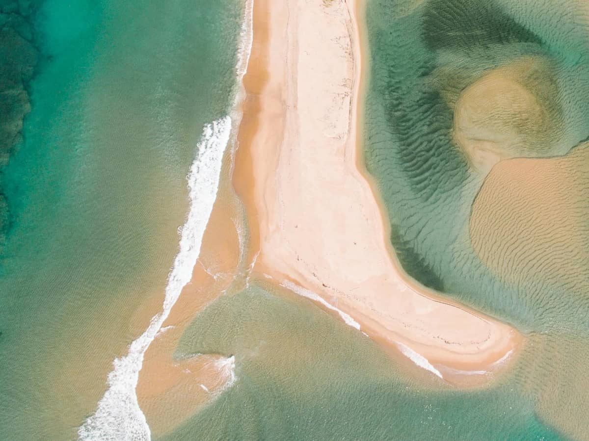 Aerial view of a sandbar splitting turquoise waters, showing waves meeting from two directions at a narrow tip of land.