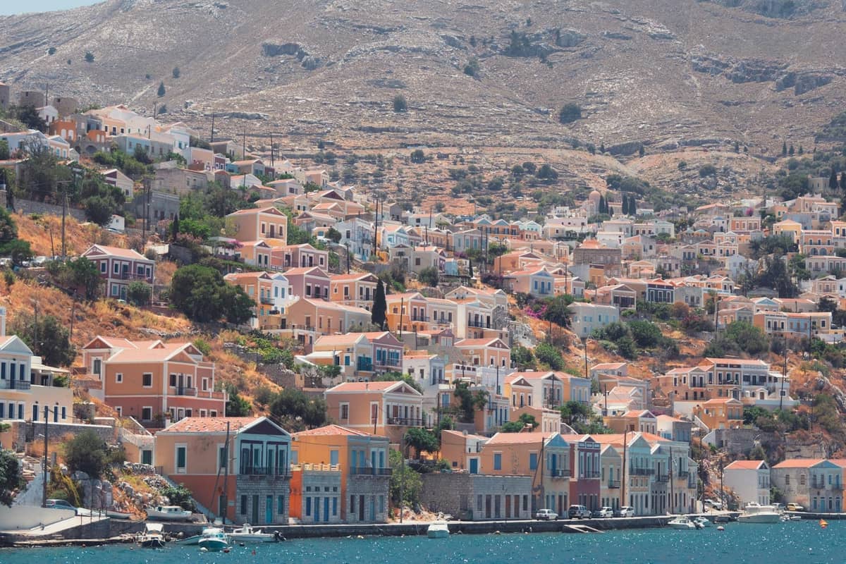 Panoramic view of Symi Island’s colorful neoclassical houses climbing up the hillside above a bright blue harbor, with arid mountains in the background.