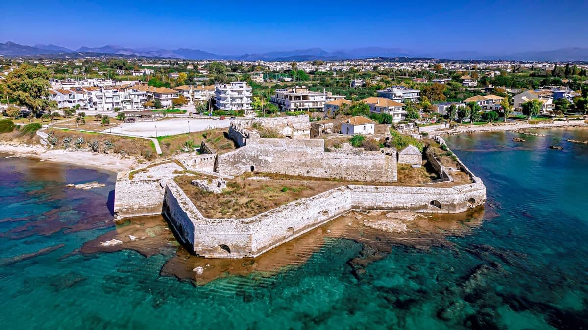 Aerial view of a fortified medieval stone structure jutting into turquoise waters with modern Rhodes town in the background.