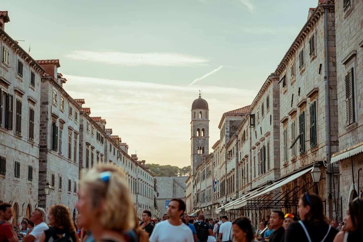 Crowds of tourists walk between historic stone buildings on a narrow street in warm sunlight, with a medieval bell tower visible in the background.