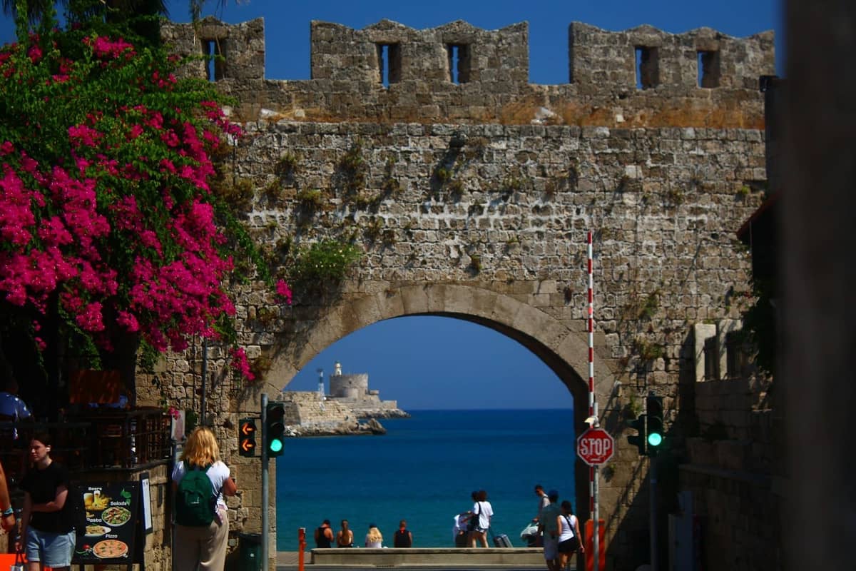 Medieval city gate with stone walls, archway opening to the sea, people walking through, and vibrant bougainvillea in Rhodes Old Town.