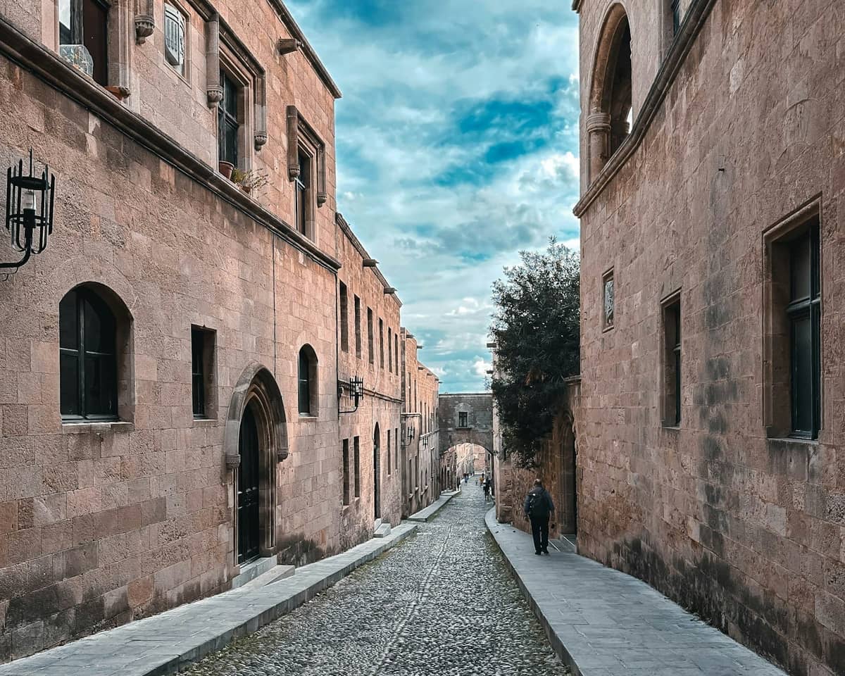 A wide cobbled street lined with medieval stone buildings in Rhodes Old Town, leading to an arched gateway under a blue sky.