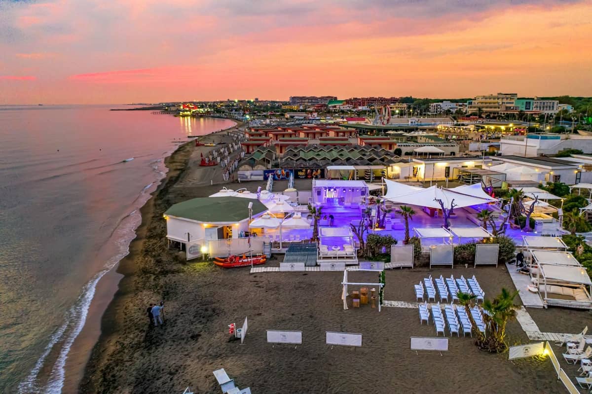 A lively beach club at sunset with purple lighting and stylish decor, located right on the sand in a bustling coastal area, evoking Rhodes nightlife.