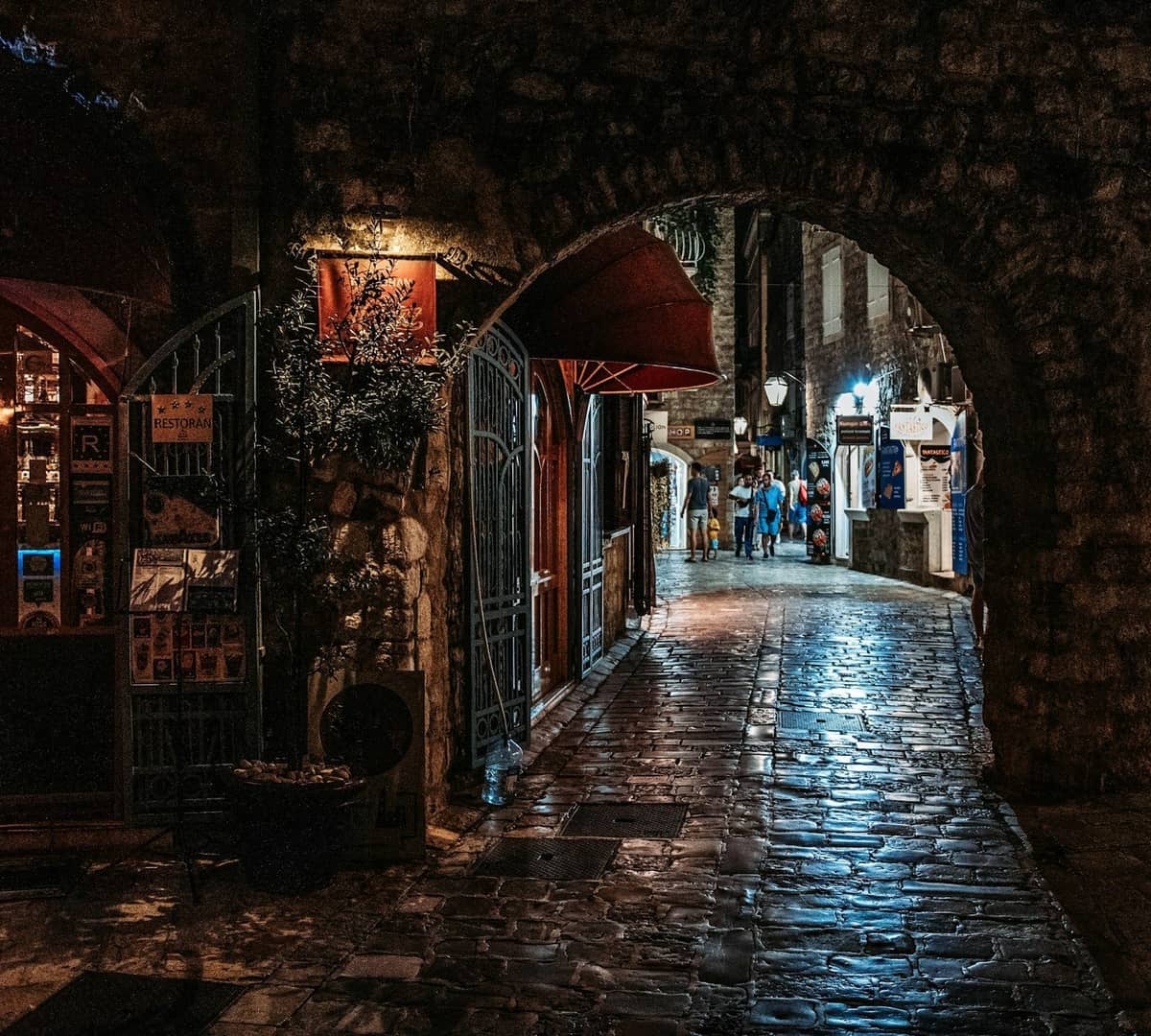 Night scene in Rhodes Old Town showing a stone archway, warmly lit bars, and people walking on a wet cobblestone street.