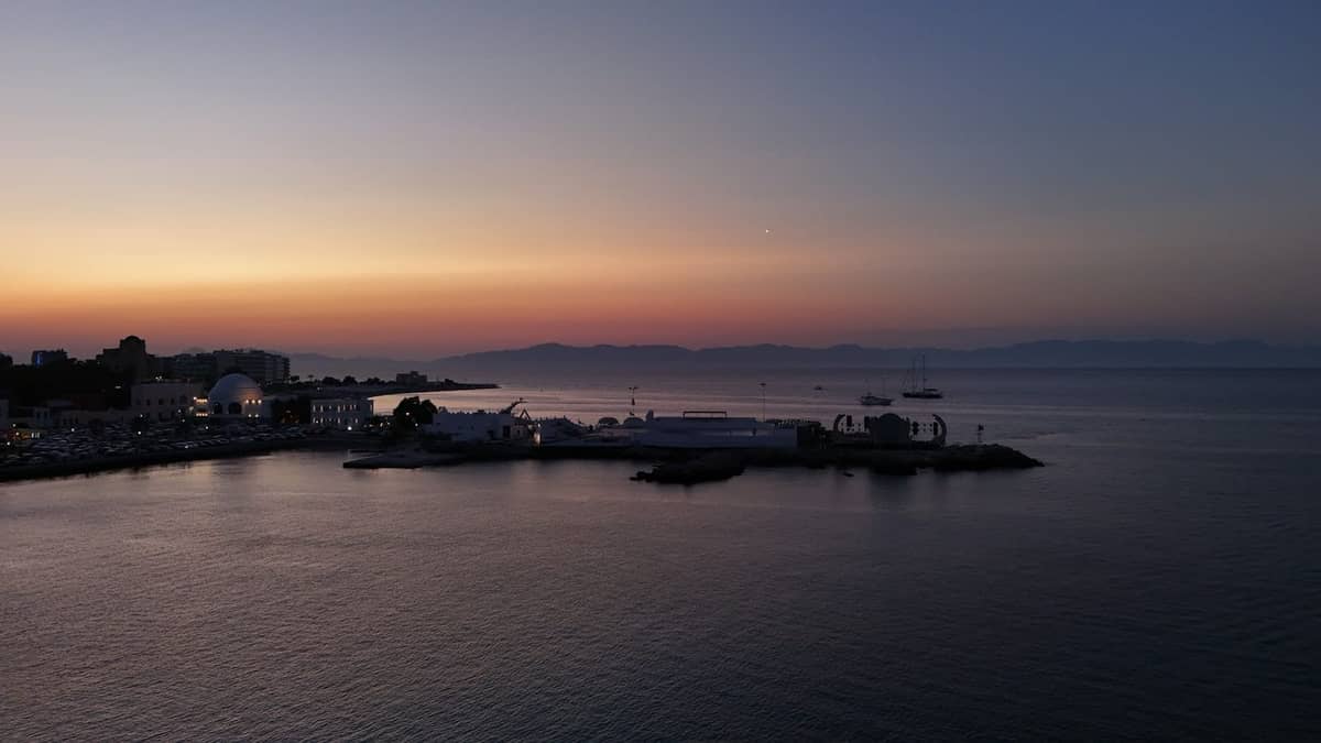 Sunset view of the waterfront in Rhodes with boats on the water, a marina, and distant mountains under a twilight sky.
