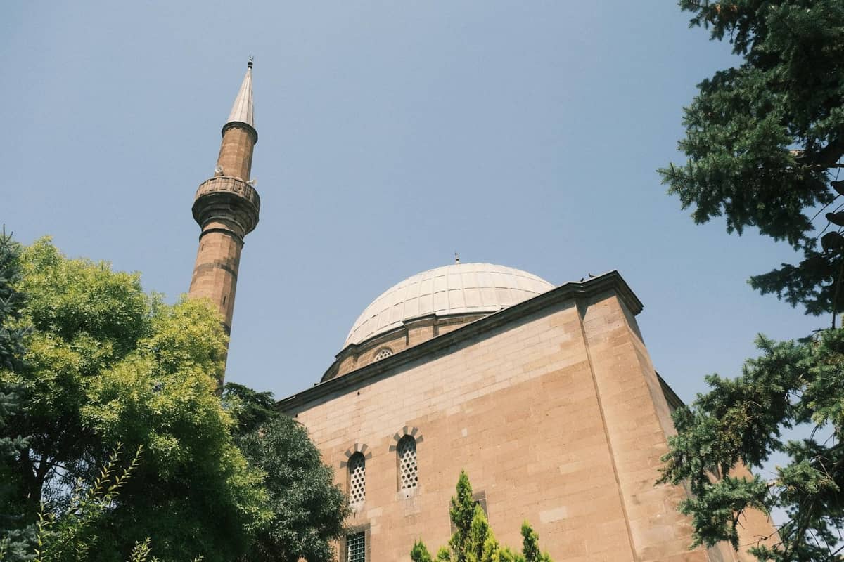 A stone mosque with a single minaret rises behind green trees under a clear sky, showing typical Ottoman architecture.