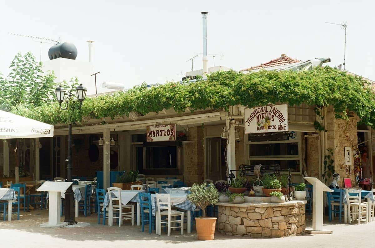 Outdoor seating at a traditional Greek taverna in Rhodes, with blue chairs, tables, and leafy vines overhead, evoking authentic local dining.