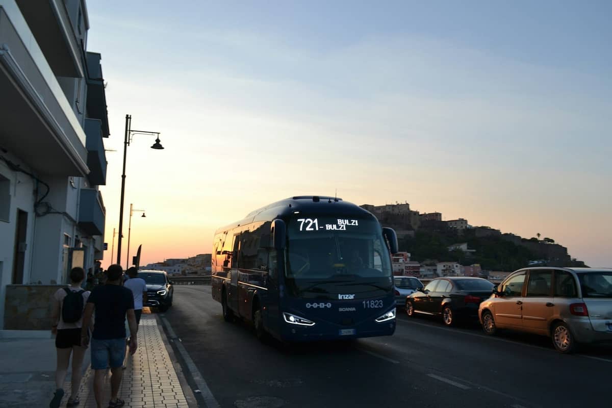 Public bus driving along a seaside road in Rhodes at sunset, with people walking on the sidewalk and a hilltop landmark in the background.