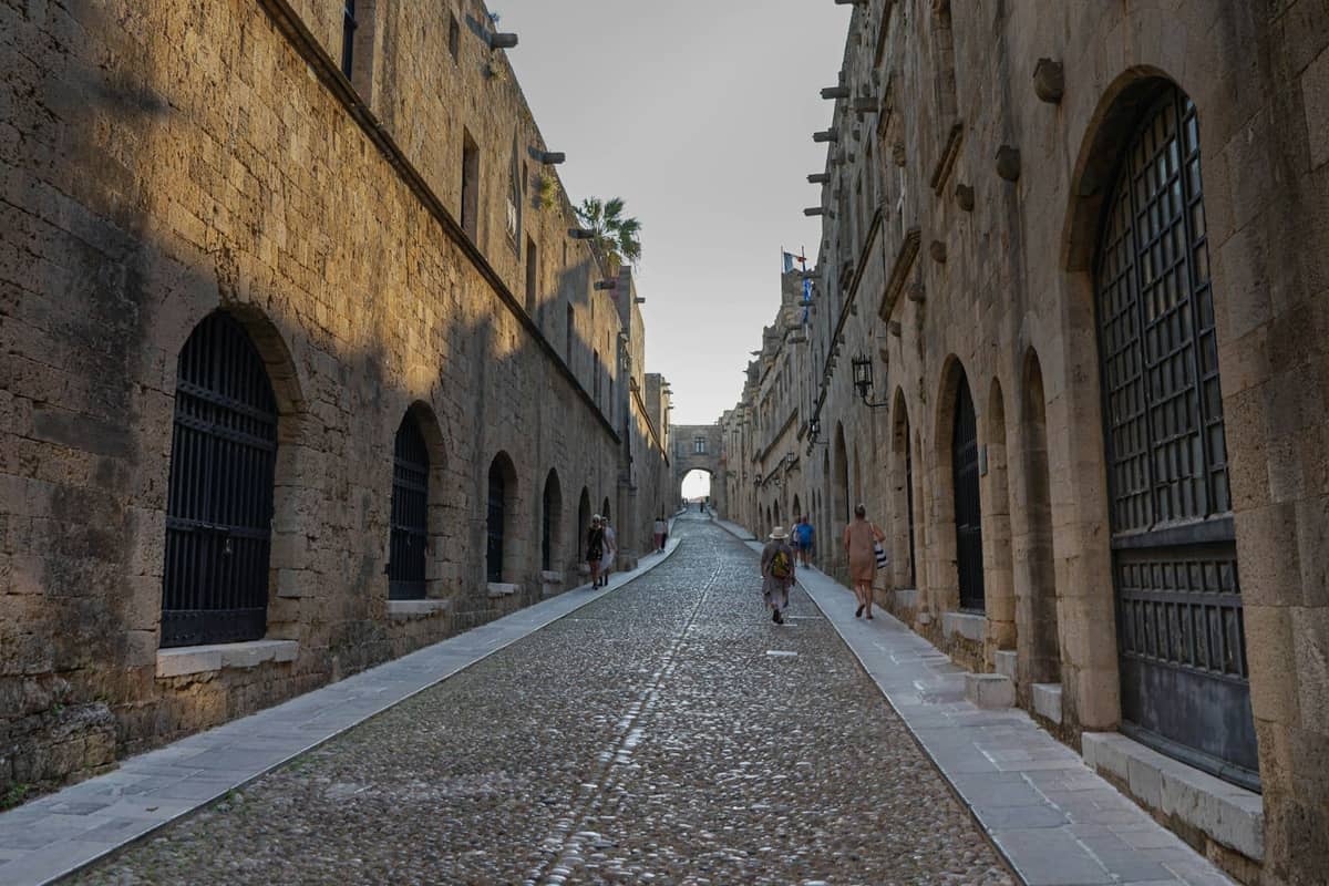 Sunlit cobblestone street flanked by medieval stone buildings in the old city of Rhodes, people strolling in the distance.