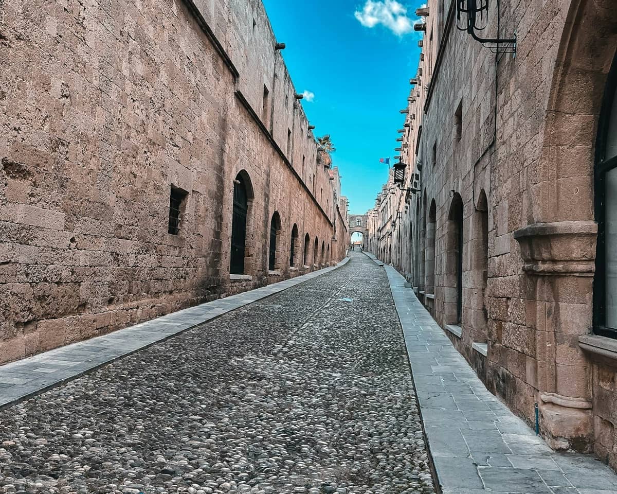 View down the medieval Street of the Knights in Rhodes Old Town, with tall stone buildings and a bright blue sky overhead.