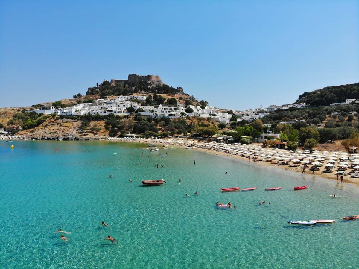 View of Lindos with turquoise bay, whitewashed houses, sandy beach, and the clifftop acropolis in the background under a clear blue sky.