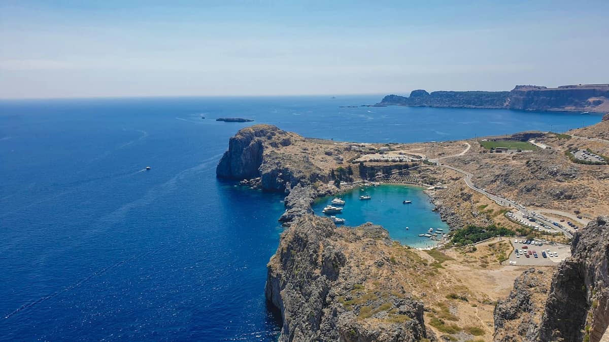 A stunning aerial view of a rocky cove with turquoise water in Rhodes, boats anchored in the bay, and rugged coastline meeting the deep blue Aegean Sea.