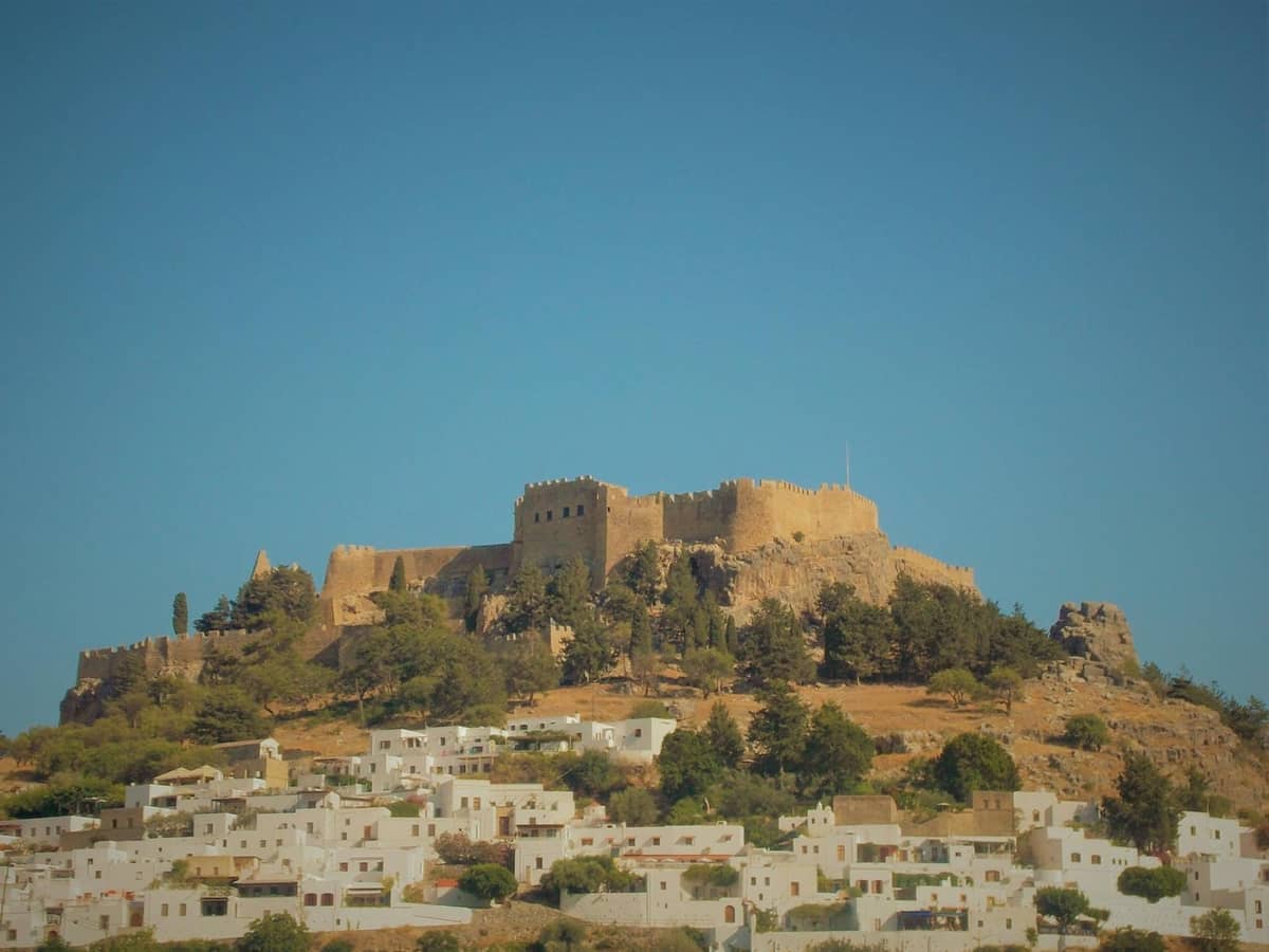 Sunlit hilltop castle overlooking white houses in Rhodes, with clear blue sky and panoramic view of the surrounding village.