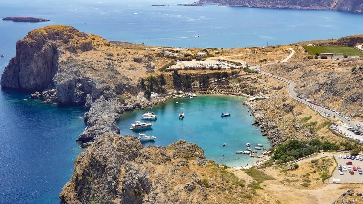Aerial view of St. Paul’s Bay in Rhodes, featuring turquoise water, boats, and rocky coastline under bright sunlight.