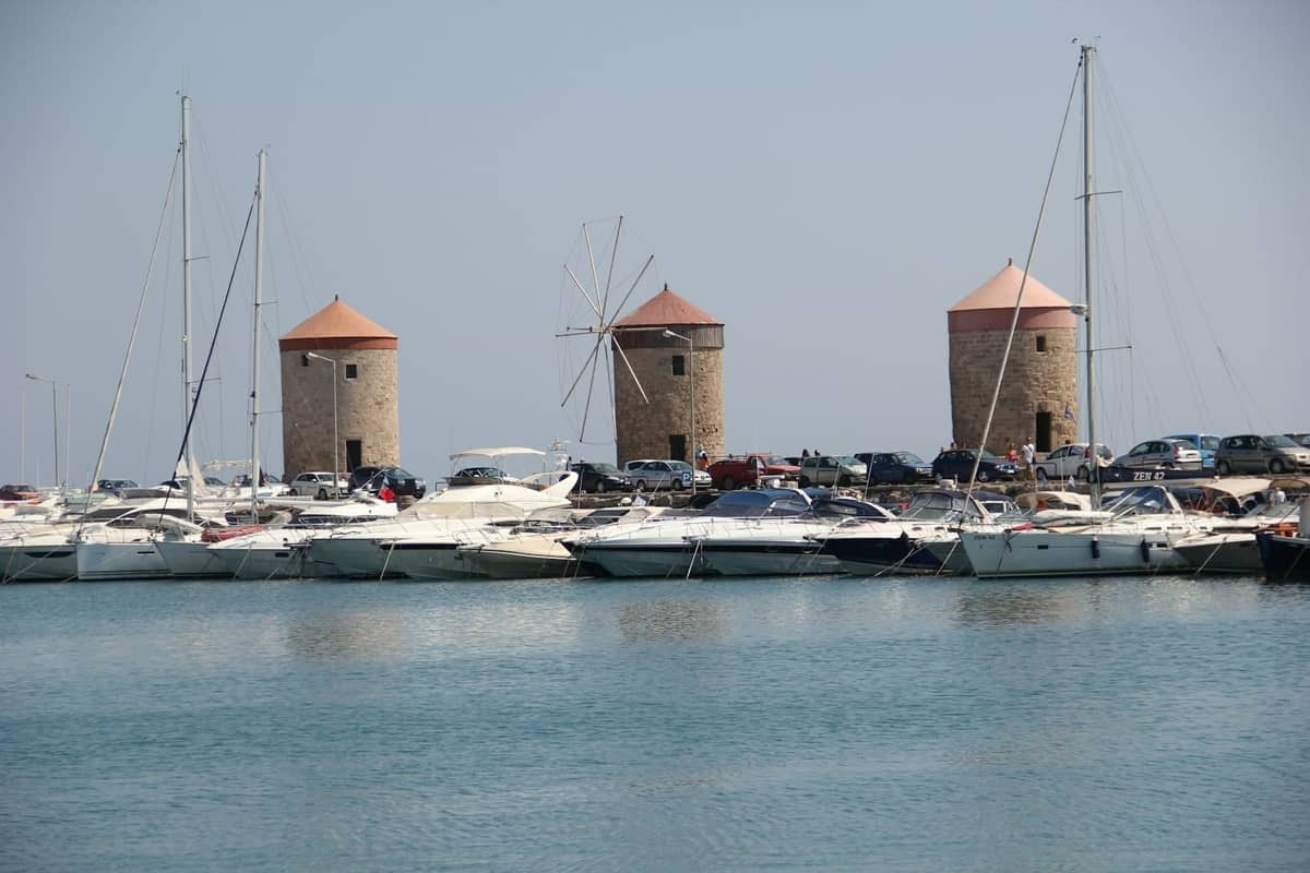 Three medieval stone windmills on the Mandraki harbor of Rhodes with yachts moored in front under a clear sky.