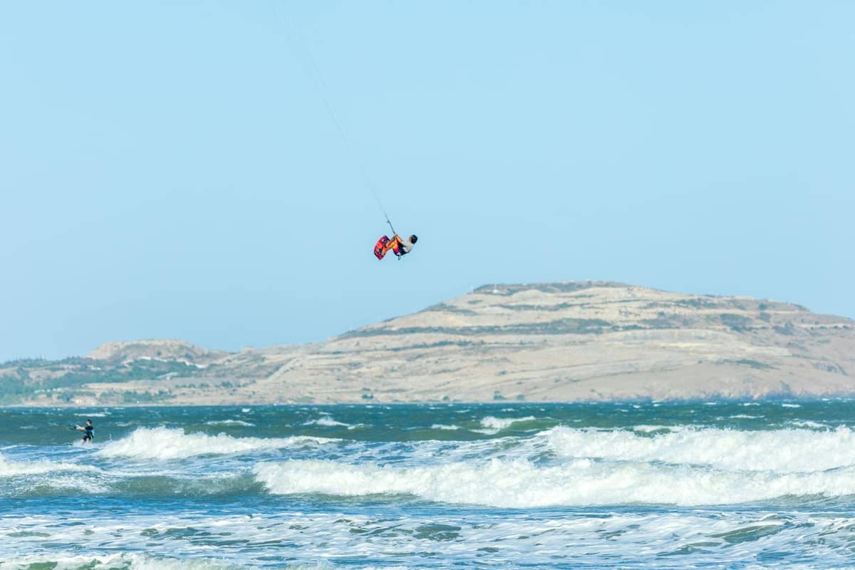 Kitesurfer riding large waves off a windy coast with dry, hilly Rhodes landscape in the background, showcasing ideal conditions for wind sports.