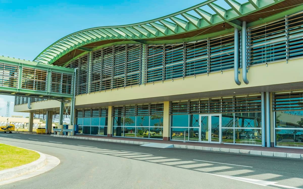 Modern exterior of an airport terminal building with curved glass and metal features under a blue sky, showing driveway and entrance area.