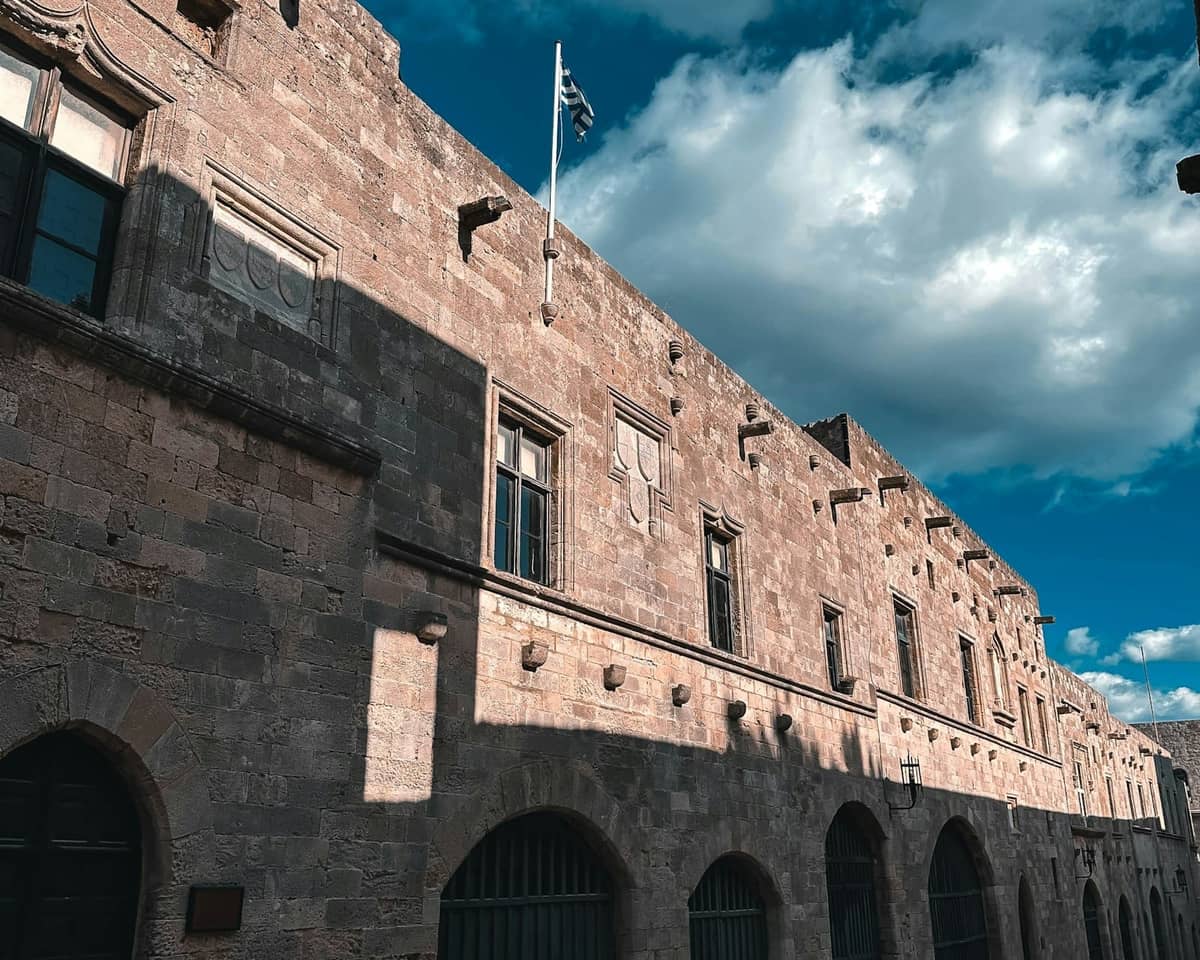 Exterior view of a historic stone building in Rhodes Old Town with arched windows, Greek flag, and dramatic sunlight and shadows.