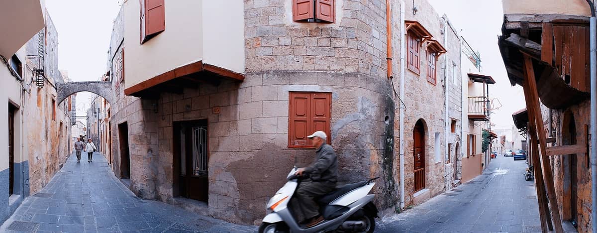 Narrow cobbled medieval street in Rhodes Old Town with stone buildings, archways, a couple walking, and a man on a scooter.