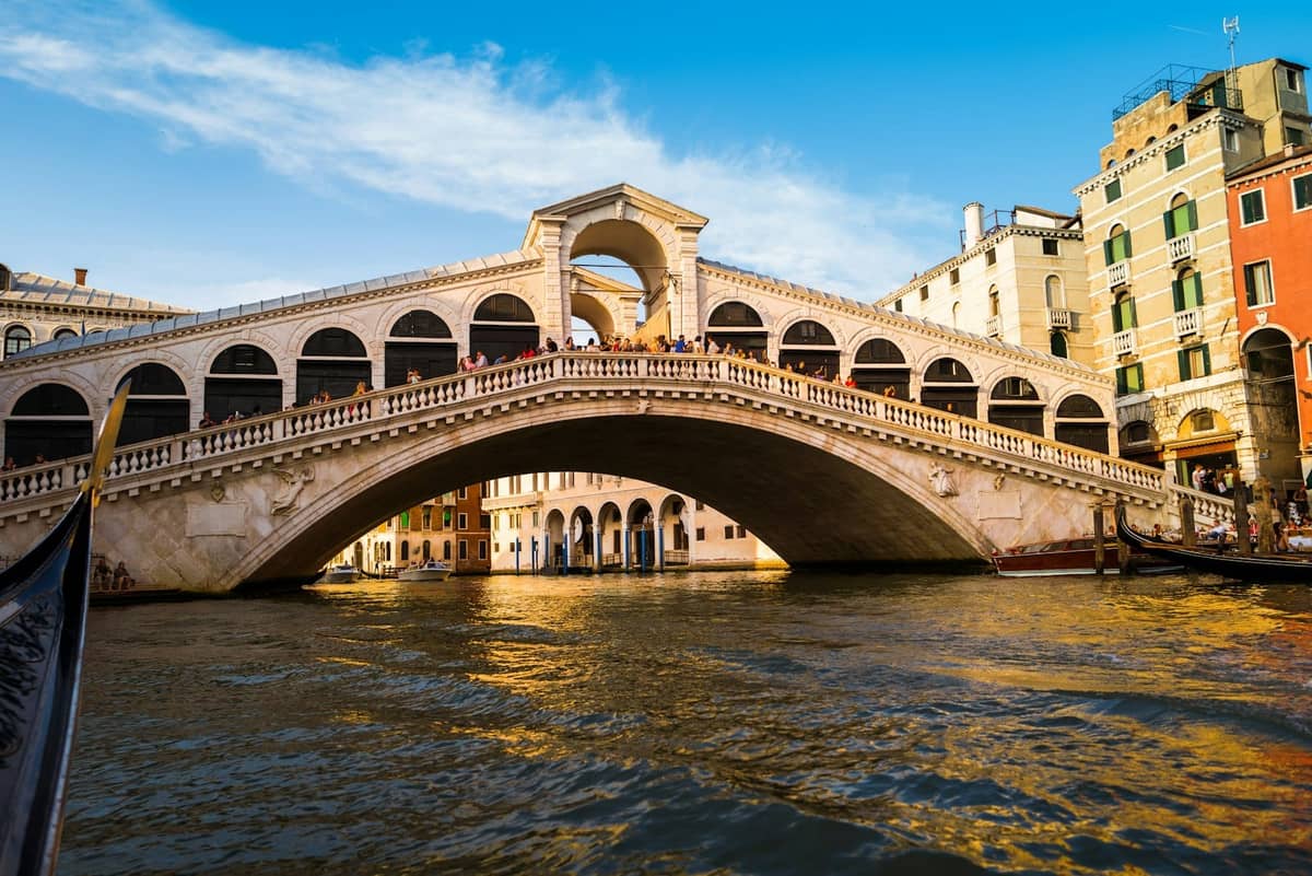 Blick auf die Rialtobrücke in Venedig, die den Canal Grande überspannt, mit historischen Gebäuden auf beiden Seiten und Spiegelungen im Wasser bei Sonnenuntergang.